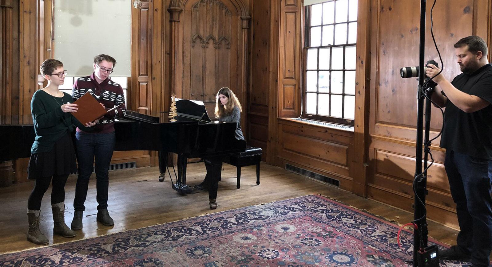 Photo of two students standing and singing, with a woman playing a grand piano behind them. 的y are being filmed by a person on the left of the photo.